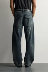 Jeans loose fit de hombre en denim de algodón color gris oscuro con efecto lavado. Presenta perneras con costuras giradas hacia los tobillos, tiro medio, cierre frontal con botones y bolsillos frontales con bolsillo cerillero.