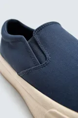 Zapatilla tipo slip-on de lona color taupe, con elástico lateral para facilitar el calce y suela de goma gruesa color crema.
