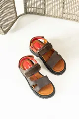 Sandalias de cuero marrón con diseño de tiras anchas y ajuste mediante velcro. Presentan una plantilla acolchada color tostado con detalles en rojo y una suela de goma negra con plataforma dentada.