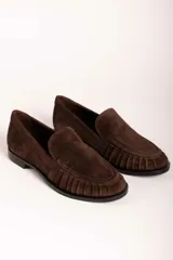 Mocasín de gamuza color marrón, con detalle de frunces en el borde superior y suela plana de goma negra.