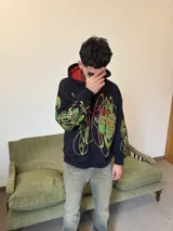 Hoodie oversize de color negro con estampado gráfico en pecho y mangas en tonos amarillos y verdes. Presenta un forro interior de capucha en color rojo con textura tejida y logo bordado en la manga.