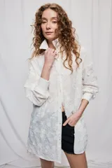Vestido camisero corto y de silueta amplia en color blanco, con cuello clásico, cierre frontal de botones y mangas largas. Cuenta con diseño bordado floral en toda la prenda y dobladillo asimétrico, más largo en la parte trasera.