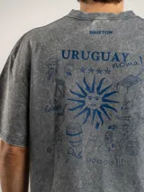 Remera oversize de algodón con efecto lavado ácido en color gris oscuro. Presenta un estampado gráfico en la espalda con motivos culturales uruguayos en color azul, incluyendo el sol de la bandera, figuras de fútbol, mate, tambor de candombe y frases típicas.