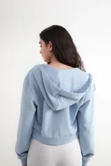 Buzo corto con capucha y cierre frontal de cremallera metálica, de manga larga y color celeste. Cuenta con bolsillos tipo canguro y puños elastizados.