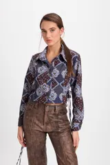 Camisa corta estilo corset con ajuste entallado, cuello clásico, cierre frontal con botones y detalle de lazo lateral. Presenta un estampado geométrico y floral en tonos azules y marrones.