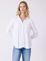 Camisa blanca de rayón con cuello solapa, escote en V y cierre frontal con botones. Presenta plisados verticales en la parte delantera y un canesú con tablón central en la espalda. El bajo es redondeado y ligeramente más largo en la espalda.