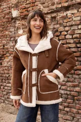 Campera estilo gamulán color tostado con interior y bordes de corderito sintético color crema. Presenta capucha amplia, cierre frontal con trabas y broches metálicos, y bolsillos delanteros grandes. Posee un calce relajado y diseño de textura suave.