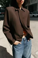 Blazer de corte estructurado en color marrón chocolate, con cuello solapa, hombros marcados y cierre frontal cruzado con botón oculto. Presenta un bolsillo superior tipo ojal y un diseño de bajo redondeado.