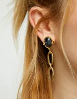 Pendientes colgantes bañados en oro 18k. Cada pendiente presenta un cristal facetado ovalado de color gris oscuro engastado, del cual cuelgan dos eslabones alargados de metal dorado.