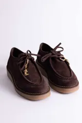 Zapato tipo mocasín acordonado de gamuza color marrón oscuro, con suela de goma color caramelo y detalle de dije metálico dorado en forma de bota colgando de los cordones.