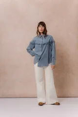 Camisa de denim liviano con calce relajado, cuello camisero clásico y cierre frontal con broches metálicos. Presenta un canesú con volado en el delantero y la espalda, mangas largas con puños abotonados y terminación inferior redondeada.