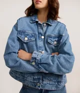 Campera de jeans de corte boxy, color azul medio lavado, con cuello, cierre frontal de botones, bolsillos con solapa y botones en el pecho, y recortes visibles.