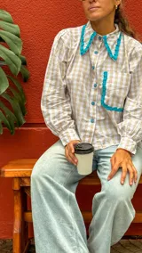 Camisa de manga larga con estampado cuadrillé en tonos beige y blanco, con cuello camisero y bolsillo frontal adornados con volados en color turquesa.