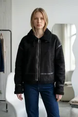 Campera tipo gamulán de color negro, confeccionada en gamuza sintética con interior y detalles en corderito negro. Presenta cierre frontal, cuello solapa y puños con terminación en corderito.