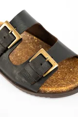Sandalias de cuero negro con diseño de doble tira con hebillas metálicas doradas, plantilla de corcho y suela plana marrón.