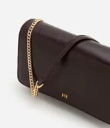 Cartera bandolera pequeña de poliuretano color marrón oscuro con acabado texturizado. Cuenta con solapa frontal, logo metálico dorado en la parte inferior y correa larga de cadena metálica dorada.