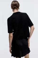 Saco de punto suave color negro, de manga corta y cuello redondo. Presenta cierre frontal con botones forrados y una moña decorativa de organza en la parte superior.