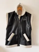 Chaleco negro de cuerina con interior de corderito sintético, bolsillos frontales y hebillas decorativas.