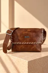 Bandolera de cuero color marfil, modelo Teka de Pecarí, con costuras cruzadas en hilo marrón oscuro en los bordes de la solapa y placa metálica frontal con la marca grabada. Incluye correa ajustable para el hombro.