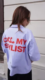 Camisa de corte boxy fit color celeste, con mangas cortas y cuello tipo polo. Presenta un estampado frontal en color rojo con la frase 'CALL MY STYLIST' y un detalle bordado en color negro con la firma 'FACHAPOSTA'.