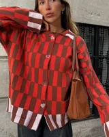 Saco tejido de punto con diseño de cuadros en tonos rojo, marrón y rosa claro. Presenta corte recto oversize, cierre frontal con botones en forma de corazón y dos bolsillos delanteros.