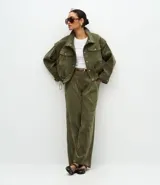 Campera de pana cotelé color verde militar, con cuello camisero, cierre frontal con botones metálicos y bolsillos frontales con solapa. Presenta diseño de sisa desplazada y cordones ajustables en el ruedo.