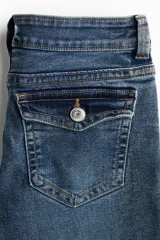Jean de denim elástico color azul con efecto deslavado, tiro bajo, corte ajustado hasta la rodilla y diseño acampanado. Cuenta con cierre de botón y cremallera, bolsillos delanteros y bolsillos traseros con solapa y botón.