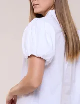 Camisa blanca de calce recto confeccionada en algodón elástico, con cuello mao, abertura frontal en V y cartera con botones ocultos. Presenta mangas cortas abullonadas con puños elastizados y ruedo redondeado.