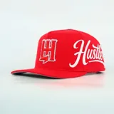 Gorra roja con visera plana, bordado frontal con iniciales rodeadas de pedrería y texto lateral bordado en color blanco.