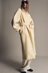 Abrigo largo color crema confeccionado en mezcla de lana. Diseño oversize con cuello en V, mangas largas y cinturón ajustable con lazada.