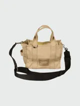 Bolso tipo tote de lona color beige de la marca Marc Jacobs. Presenta el texto 'THE TOTE BAG' estampado en el frente en color blanco, dos asas de mano superiores y una correa de hombro ajustable y removible en color negro.