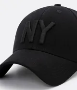 Gorra de seis paneles con ala curva, confeccionada en algodón color marrón. Presenta un bordado frontal con las letras NY en color blanco y cuenta con cierre strapback ajustable en la parte posterior.