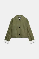 Cazadora verde militar de corte crop, confeccionada en hilatura con lino, con cuello solapa, manga larga y bolsillos de plastrón en el delantero. Cuenta con cierre frontal de botones oscuros y detalles a rayas en contraste en los puños y el interior.