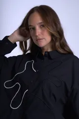 Camisa negra de manga larga con cuello clásico y cierre frontal con botones. Presenta bordados abstractos en blanco y negro en el frente.