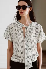 Blusa de viscosa con estampado de lunares, cuello redondo con escote en pico, volantes y lazada frontal. Presenta manga corta y bajo recto.