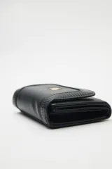 Billetera compacta de cuero sintético negro, con solapa que combina texturas lisas y efecto reptil. Incluye un aplique metálico circular con logo grabado en el frente y cierre con broche.