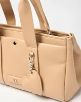 Cartera tipo tote de símil cuero color beige, con doble asa de mano, correa ajustable y desmontable, y llavero colgante en forma de corazón. Cuenta con detalle de logo metálico en el frente y broche a presión.