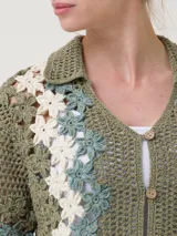 Sweater verde de tejido tipo crochet, con cuello camisero, mangas cortas y cierre frontal de botones. Presenta un diseño de bandas laterales con aplicaciones florales en tonos blancos y celestes.
