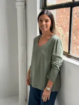 Blusa de manga larga en color verde oliva, con cuello en V y costuras decorativas visibles. Presenta un corte recto y holgado con un ligero fruncido en los hombros.