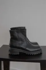 Bota corta de cuero negro con punta cuadrada, suela track y detalle de estribo desmontable con hebilla metálica.