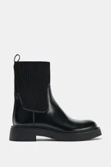 Botín de estilo urbano con suela track, confeccionado en cuero sintético negro con caña de tejido elástico acanalado. Presenta punta redonda, tirador trasero y diseño tipo pull-on.