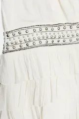 Vestido corto de gasa color blanco, con escote en V cruzado, sin mangas y diseño con volantes en el bajo. Presenta un detalle de tachas plateadas en la cintura.