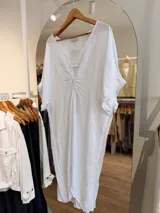 Vestido de lino blanco con silueta oversize, escote en V y detalle de nudo delantero con efecto drapeado. Cuenta con mangas amplias y diseño asimétrico, más largo en la parte trasera.