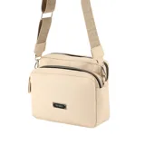 Bandolera de diseño rectangular en color beige, confeccionada en material simil cuero con textura granulada. Cuenta con dos compartimentos principales con cierres metálicos, placa metálica decorativa con el logo de la marca en el frente y correa ajustable de tela con diseño grabado.