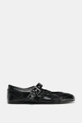 Zapato tipo bailarina de acabado acharolado en color negro, con punta redonda y suela plana. Presenta una tira ajustable con hebilla metálica y bandas elásticas cruzadas en el empeine.