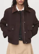 Chaqueta corta de algodón en color marrón oscuro, con cuello de efecto piel en tono contrastante. Presenta cierre frontal con cremallera y botones a presión, además de dos bolsillos frontales con solapa y cierre de botón.