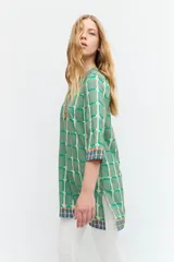 Túnica verde con estampa geométrica de cuadrados en verde más oscuro y blanco. Tiene escote en V con ribete marrón y mangas tres cuartos con ribete geométrico en puños y ruedo.