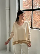 Sweater de punto color crudo con cuello en V, mangas largas y diseño holgado. Presenta detalles de franjas horizontales en color beige en los puños y en el ruedo inferior.