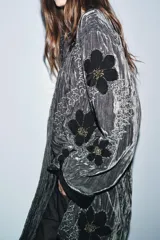 Kimono de terciopelo gris con acabado texturizado, cuello redondo y manga larga. Presenta bordados florales en hilo negro y metalizado, bolsillos delanteros y cierre frontal.