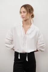 Camisa de color crudo con diseño romántico, cuello redondo con cordón ajustable terminado en borlas, y mangas tres cuartos abullonadas. Presenta detalles de bordados calados verticales en el frente y horizontales en las mangas.